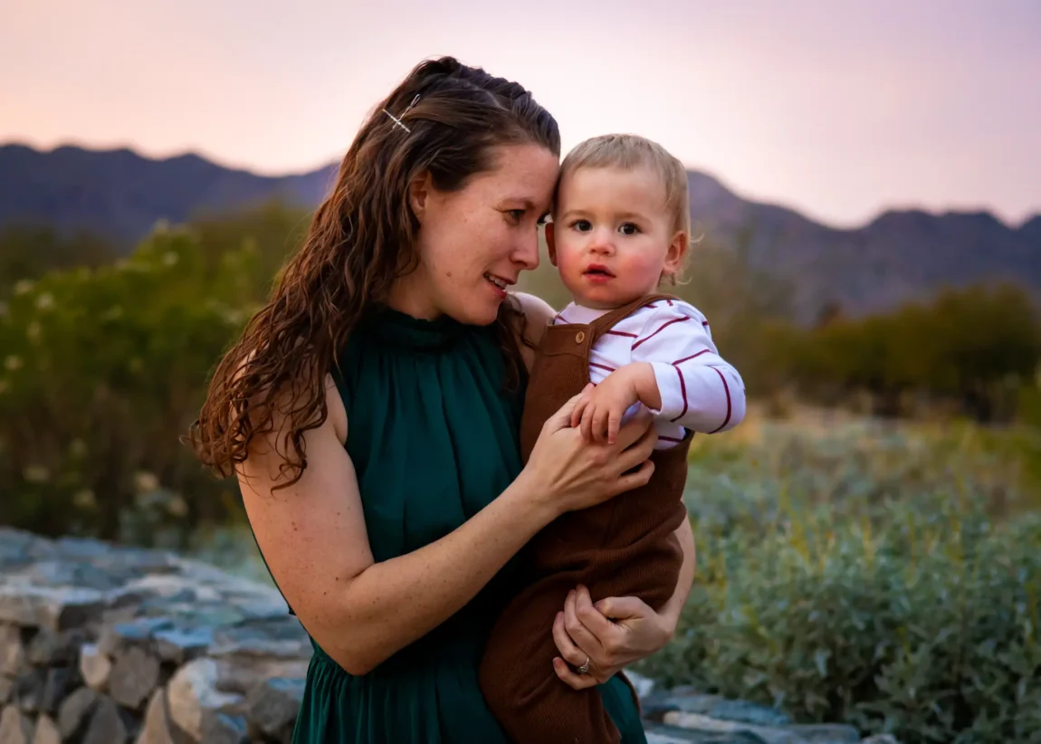 Mom in green dress holding her toddler boy cheek to cheek against an Arizona desert sunset