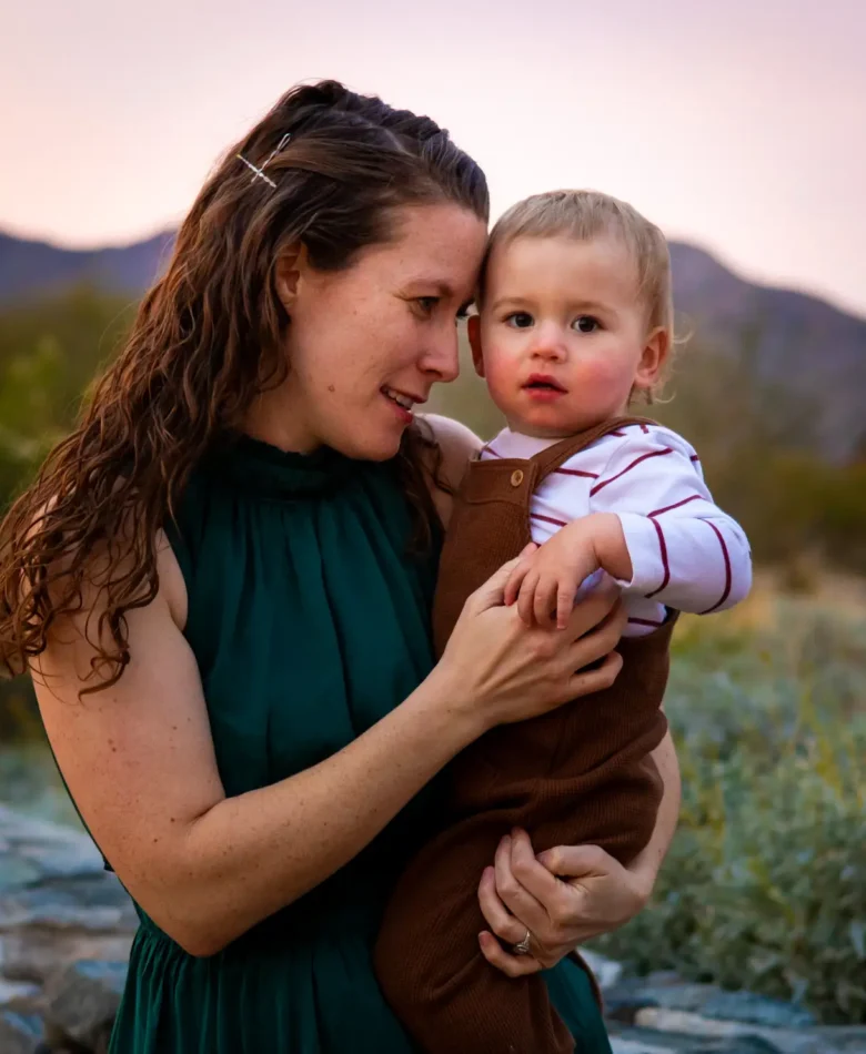 Mom in green dress holding her toddler boy cheek to cheek against an Arizona desert sunset