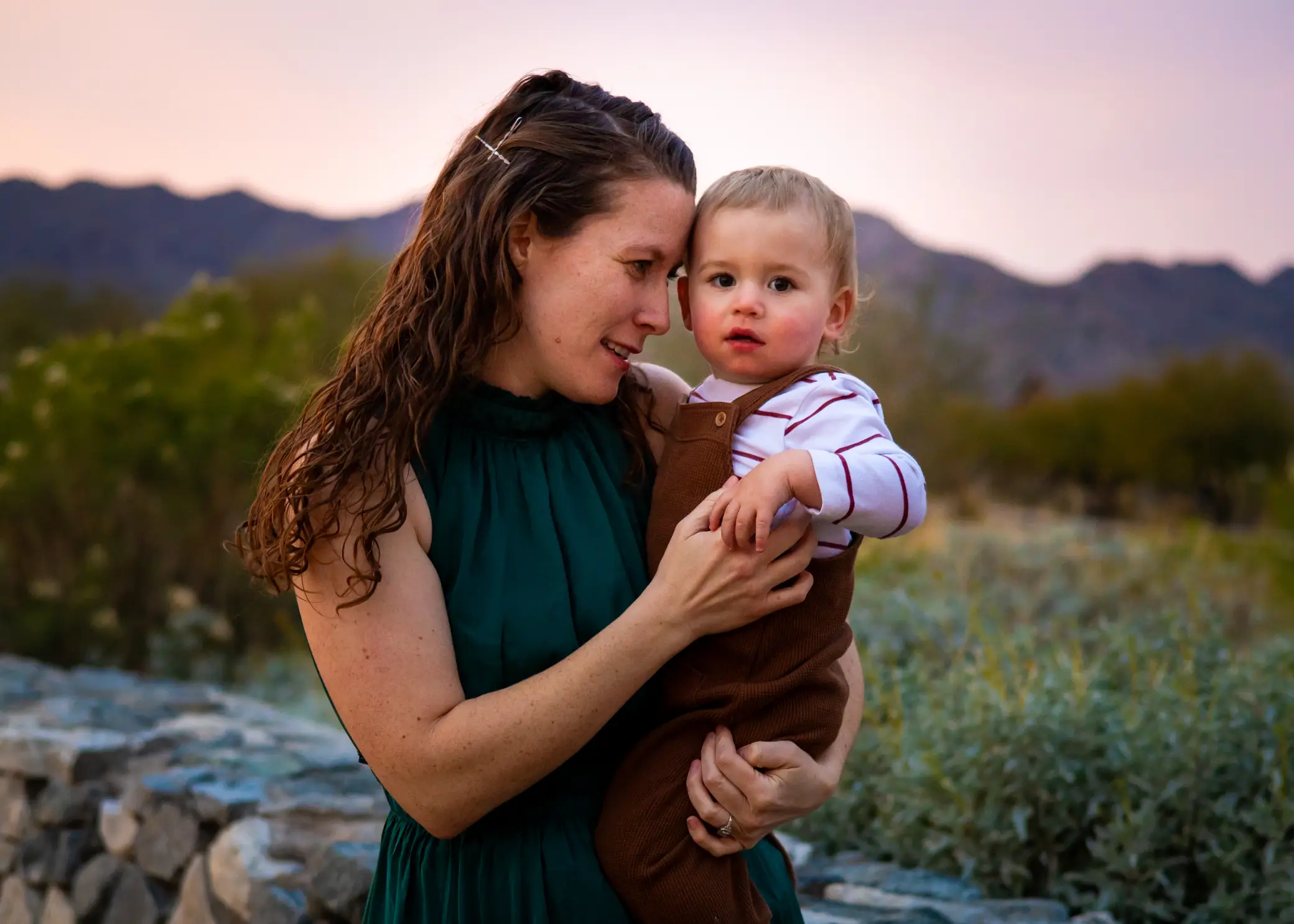 Mom in green dress holding her toddler boy cheek to cheek against an Arizona desert sunset