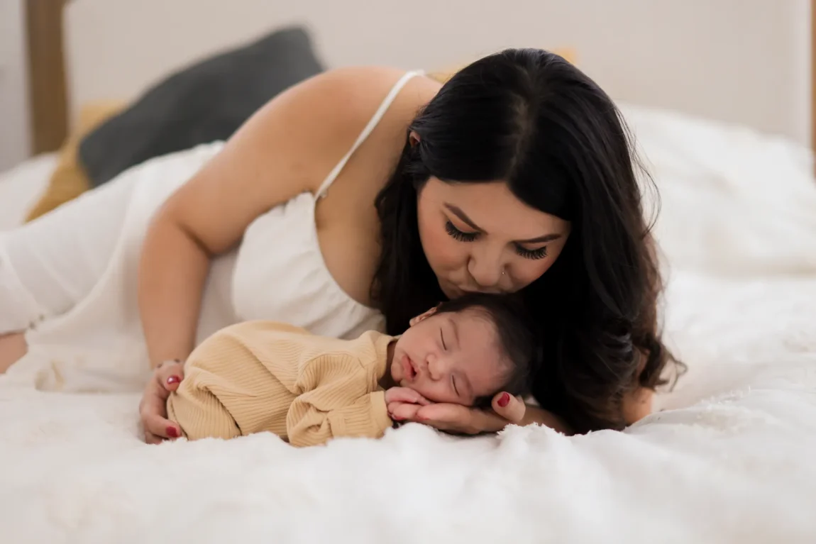 Mom gently kissing her sleeping newborn on the bed during an in-home newborn photography session