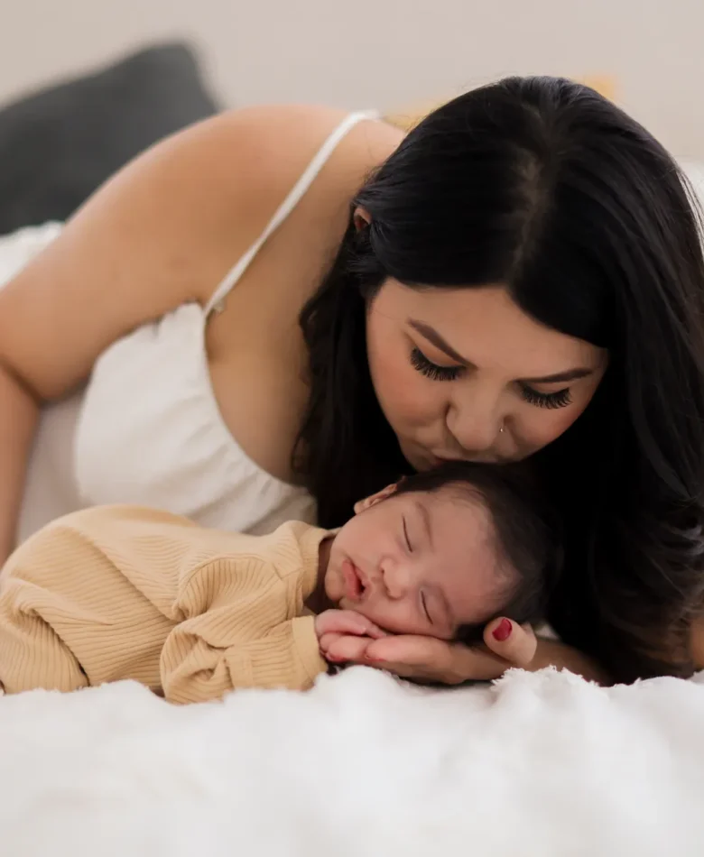 Mom gently kissing her sleeping newborn on the bed during an in-home newborn photography session