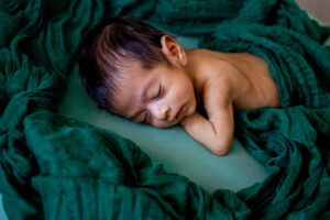 Sleeping newborn baby curled up on a teal pillow wrapped in a deep green muslin blanket during a newborn photography session in Phoenix West Valley