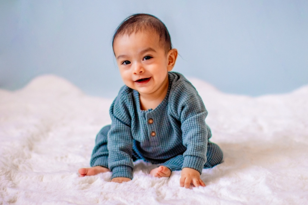 Smiling baby in a sage green waffle knit romper sitting up on a white fluffy rug during a sitter milestone session in Phoenix West Valley